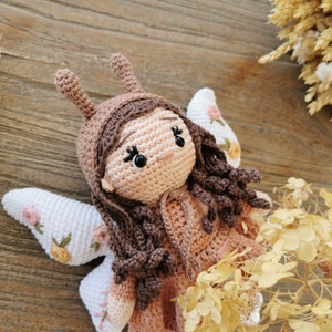 May include: A handmade crochet doll in the shape of a fairy. The doll has a light brown face, brown hair, and a matching hat with bunny ears. The doll is wearing a pink dress and has white wings with pink floral embroidery. The doll is lying on a wooden surface.