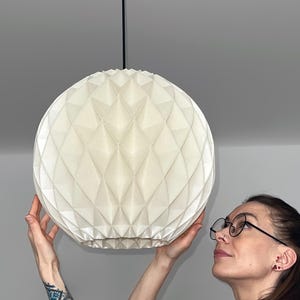 Może przedstawiać: Sferyczna lampa wisząca z białym, składanym, geometrycznym papierowym kloszem o fakturze w romby. Klosz jest zawieszony na suficie za pomocą cienkiego czarnego przewodu przymocowanego do pasującej czarnej okrągłej podsufitki.