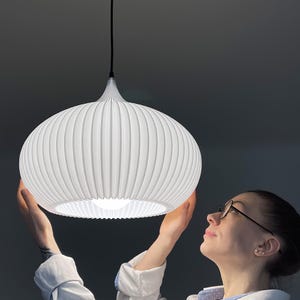May include: A white pendant light fixture with a rounded, ribbed, and pleated lampshade design. The light hangs from a black cord attached to a black ceiling canopy. The shade features a wide, circular opening at the bottom, revealing a bright light bulb.