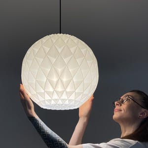 Może przedstawiać: Sferyczna lampa wisząca o białej, geometrycznej i fasetowanej powierzchni. Lampa zawieszona jest na cienkim czarnym przewodzie przymocowanym do okrągłej czarnej podsufitki, emitując z wnętrza miękkie, rozproszone światło.