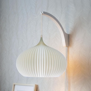 Op de afbeelding: Een witte, geplooide, ui-vormige hanglampenkap opgehangen aan een gebogen witte wandarm. De lamp heeft een verticaal geribbelde textuur en is verbonden met een wit stroomsnoer dat langs de muur naar beneden loopt.