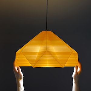 May include: A person holds up a yellow origami-style pendant light with a black cord.