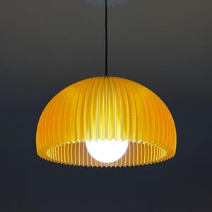 Könnte beinhalten: Gelbe, kuppelförmige Pendelleuchte. Der Lampenschirm hat ein geripptes Design und eine kugelförmige Glühbirne ist unten sichtbar. Die Leuchte hängt an einem schwarzen Kabel und einer schwarzen Deckenhalterung.