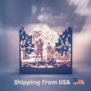 Könnte beinhalten: Eine weiße Papierkunst-Skulptur, die ein Paar zeigt, das unter einem Baum mit einem Herz steht, auf dem "Ich liebe dich" steht. Das Paar ist von Blumen und einem Teich mit Schwänen umgeben. Die Skulptur wird von hinten beleuchtet und erzeugt so ein warmes Leuchten.