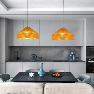 May include: Two modern pendant lights with geometric, wooden shades hang over a kitchen island with a dark countertop. The lights are suspended from black cords and have a warm, yellow glow.
