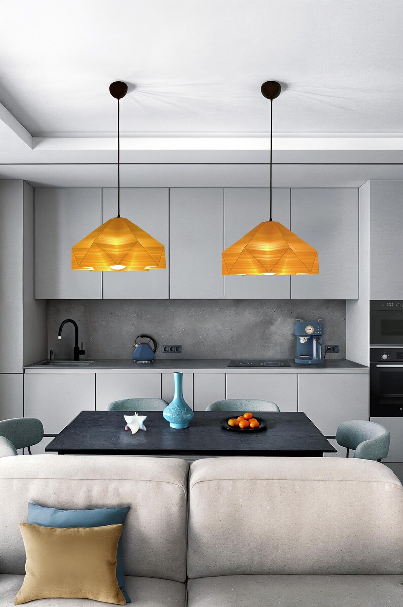 May include: Two modern pendant lights with geometric, wooden shades hang over a kitchen island with a dark countertop. The lights are suspended from black cords and have a warm, yellow glow.