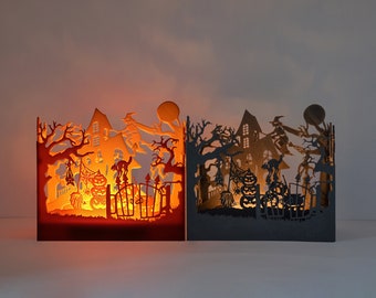 May include: Two black metal Halloween candle holders featuring a spooky silhouette design. The left holder is lit with a candle, casting an orange glow. The design includes a haunted house, a witch flying on a broomstick, a cat, and pumpkins.