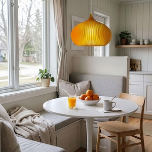 Peut inclure: Une suspension jaune vif avec un abat-jour arrondi et nervur&eacute; est suspendue au-dessus d'une table ronde &agrave; pied central blanc. La table est garnie d'un verre de jus d'orange, d'un bol d'oranges et d'une tasse en c&eacute;ramique, pr&egrave;s d'une banquette grise.