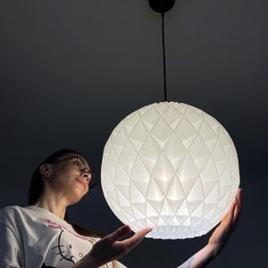 Może przedstawiać: Sferyczna lampa wisząca o białej, geometrycznej, fasetowanej powierzchni. Oprawa oświetleniowa zawieszona jest na cienkim czarnym przewodzie przymocowanym do okrągłej czarnej podsufitki, emitując miękkie, rozproszone światło z wnętrza.