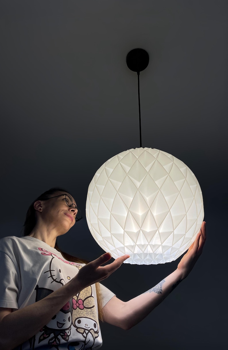 Może przedstawiać: Sferyczna lampa wisząca o białej, geometrycznej, fasetowanej powierzchni. Oprawa oświetleniowa zawieszona jest na cienkim czarnym przewodzie przymocowanym do okrągłej czarnej podsufitki, emitując miękkie, rozproszone światło z wnętrza.