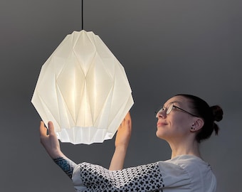 May include: A white pendant lamp with a geometric, faceted, origami-inspired shade. The light fixture features a structured, multi-sided design that emits a soft, diffused glow. It is suspended from a thin black cord attached to a black ceiling mount.