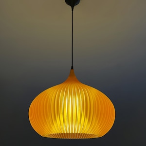 Peut inclure: Une suspension lumineuse jaune vif avec une forme arrondie, nervur&eacute;e et semblable &agrave; une citrouille. La lampe est suspendue au plafond par un fin cordon noir fix&eacute; &agrave; une petite rosace circulaire noire, diffusant une lumi&egrave;re chaude et &eacute;clatante.