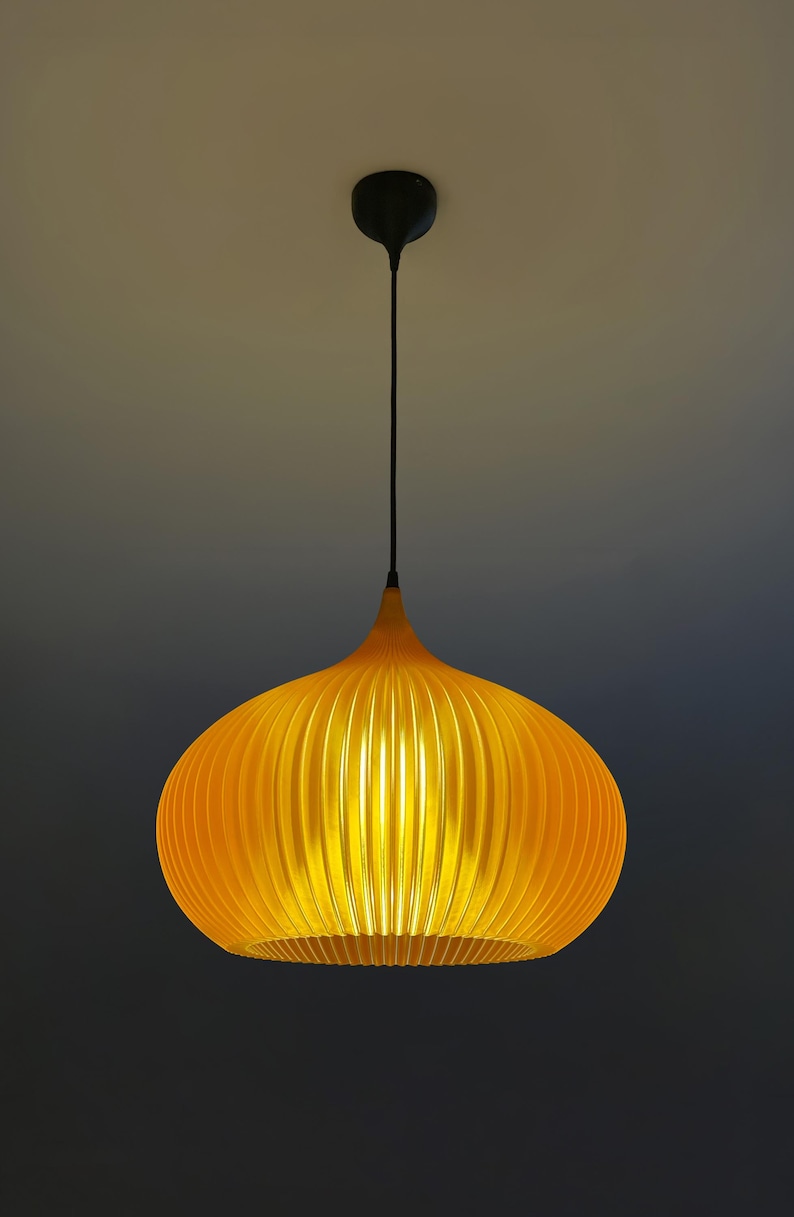 Peut inclure: Une suspension lumineuse jaune vif avec une forme arrondie, nervur&eacute;e et semblable &agrave; une citrouille. La lampe est suspendue au plafond par un fin cordon noir fix&eacute; &agrave; une petite rosace circulaire noire, diffusant une lumi&egrave;re chaude et &eacute;clatante.