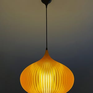 Peut inclure: Une suspension jaune vif avec un abat-jour nervur&eacute; en forme d'oignon. Le luminaire est suspendu par un fin cordon noir attach&eacute; &agrave; une petite rosace de plafond noire. La lumi&egrave;re &eacute;met un &eacute;clat chaud, soulignant les nervures verticales de l'abat-jour.