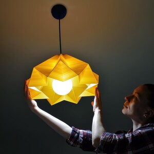 May include: A person holds up a yellow origami-style pendant light fixture. The light is on and illuminates the room.