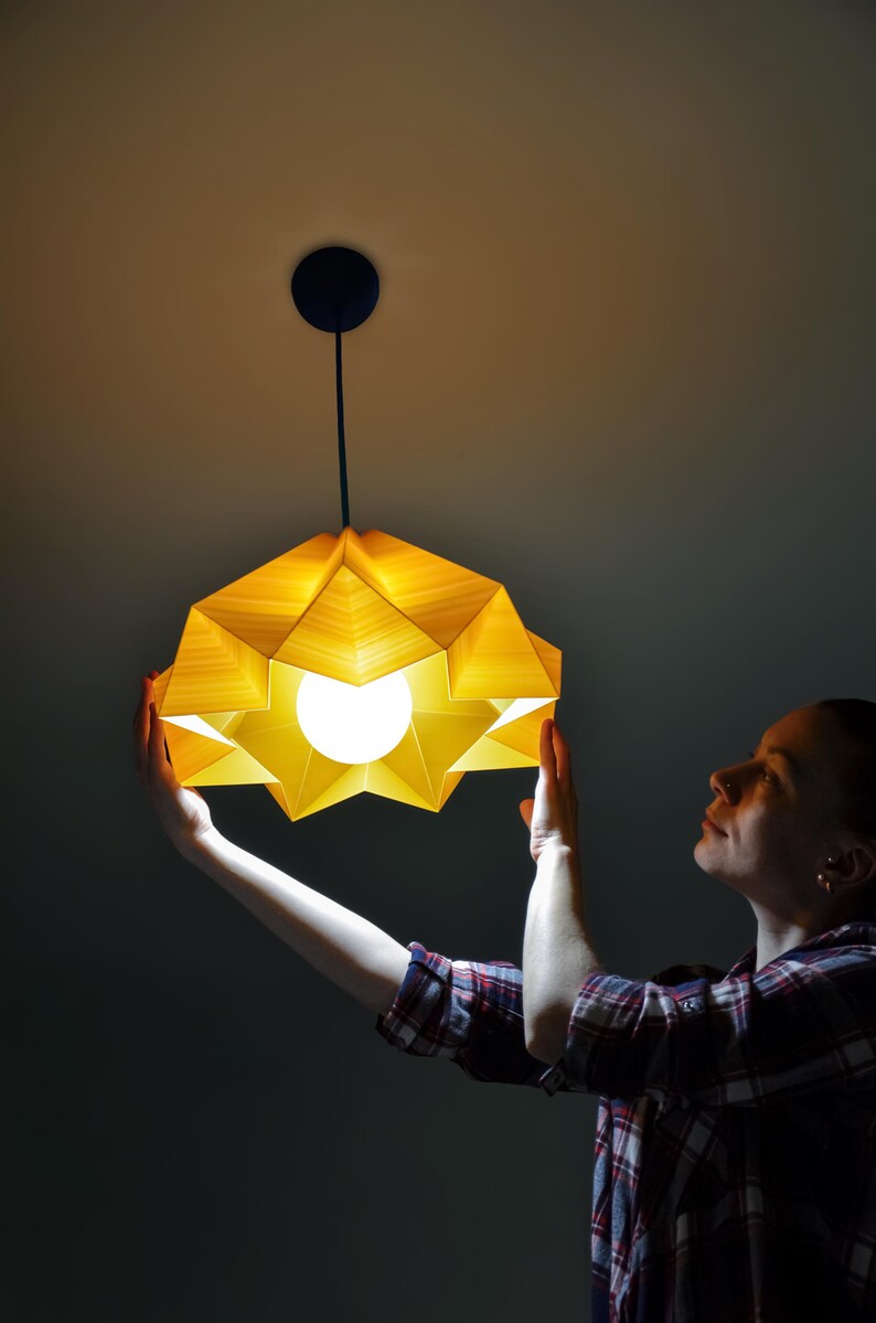 May include: A person holds up a yellow origami-style pendant light fixture. The light is on and illuminates the room.