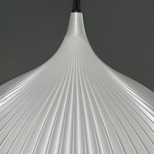 A white pendant light shade with a conical, fluted design featuring vertical ridges radiating from the center. The shade is suspended by a black cord and a black cylindrical fixture at the top, creating a clean, symmetrical appearance.