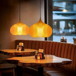 Peut inclure: Deux suspensions de couleur ambre avec un design nervur&eacute; en forme d'oignon, suspendues par des cordons noirs. Les lampes diffusent une lumi&egrave;re chaude &agrave; travers les lattes verticales, cr&eacute;ant un &eacute;clairage doux soulignant la structure courbe.