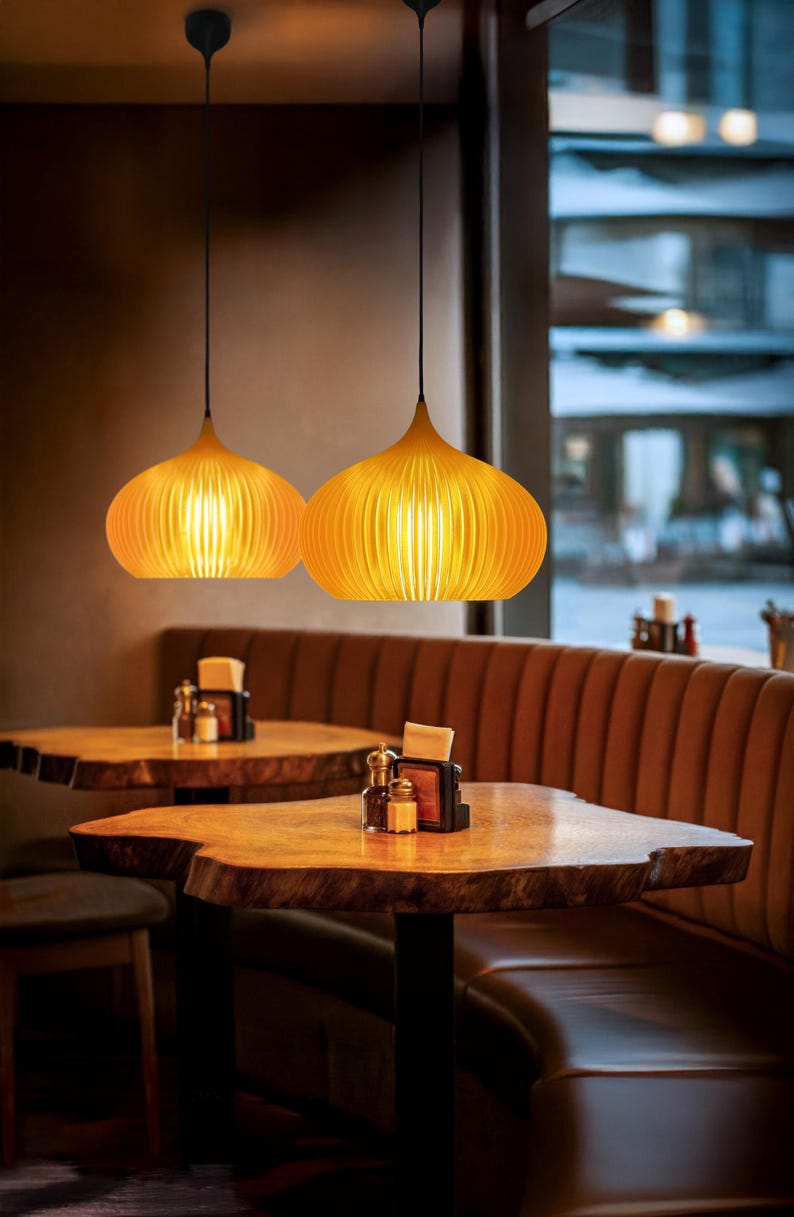 Peut inclure: Deux suspensions de couleur ambre avec un design nervur&eacute; en forme d'oignon, suspendues par des cordons noirs. Les lampes diffusent une lumi&egrave;re chaude &agrave; travers les lattes verticales, cr&eacute;ant un &eacute;clairage doux soulignant la structure courbe.