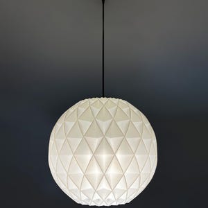 Może przedstawiać: Sferyczna lampa wisząca o białej, geometrycznej, fasetowanej powierzchni. Klosz zawieszony jest na cienkim czarnym przewodzie przymocowanym do małej, okrągłej czarnej podsufitki, emitując miękkie, rozproszone światło.