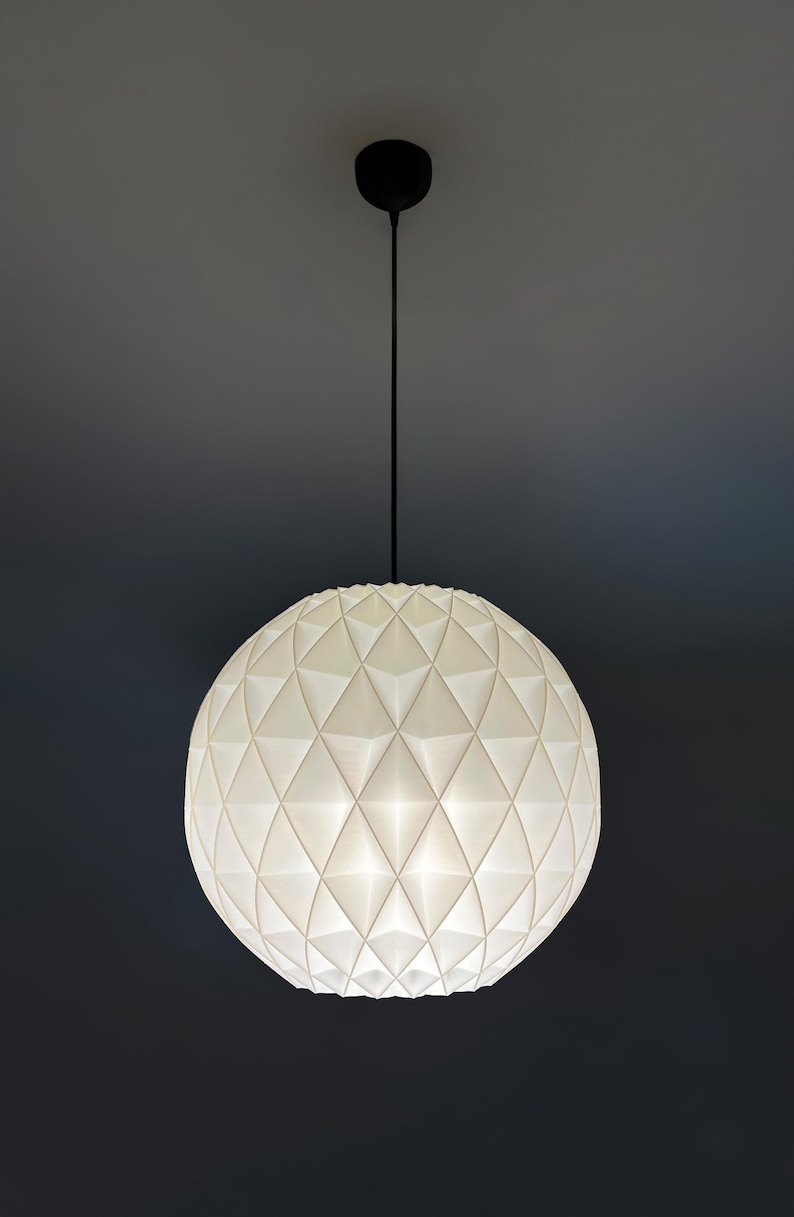 Może przedstawiać: Sferyczna lampa wisząca o białej, geometrycznej, fasetowanej powierzchni. Klosz zawieszony jest na cienkim czarnym przewodzie przymocowanym do małej, okrągłej czarnej podsufitki, emitując miękkie, rozproszone światło.