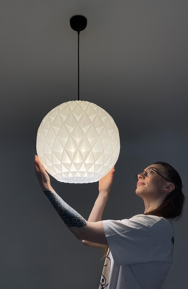 Może przedstawiać: Sferyczna lampa wisząca o białej, geometrycznej i fasetowanej powierzchni. Lampa zawieszona jest na cienkim czarnym przewodzie przymocowanym do okrągłej czarnej podsufitki, emitując z wnętrza miękkie, rozproszone światło.