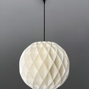 Może przedstawiać: Sferyczna lampa wisząca z białym, składanym, geometrycznym wzorem plastra miodu. Klosz jest zawieszony na cienkim czarnym przewodzie przymocowanym do małej, okrągłej czarnej podsufitki na szczycie całej konstrukcji.