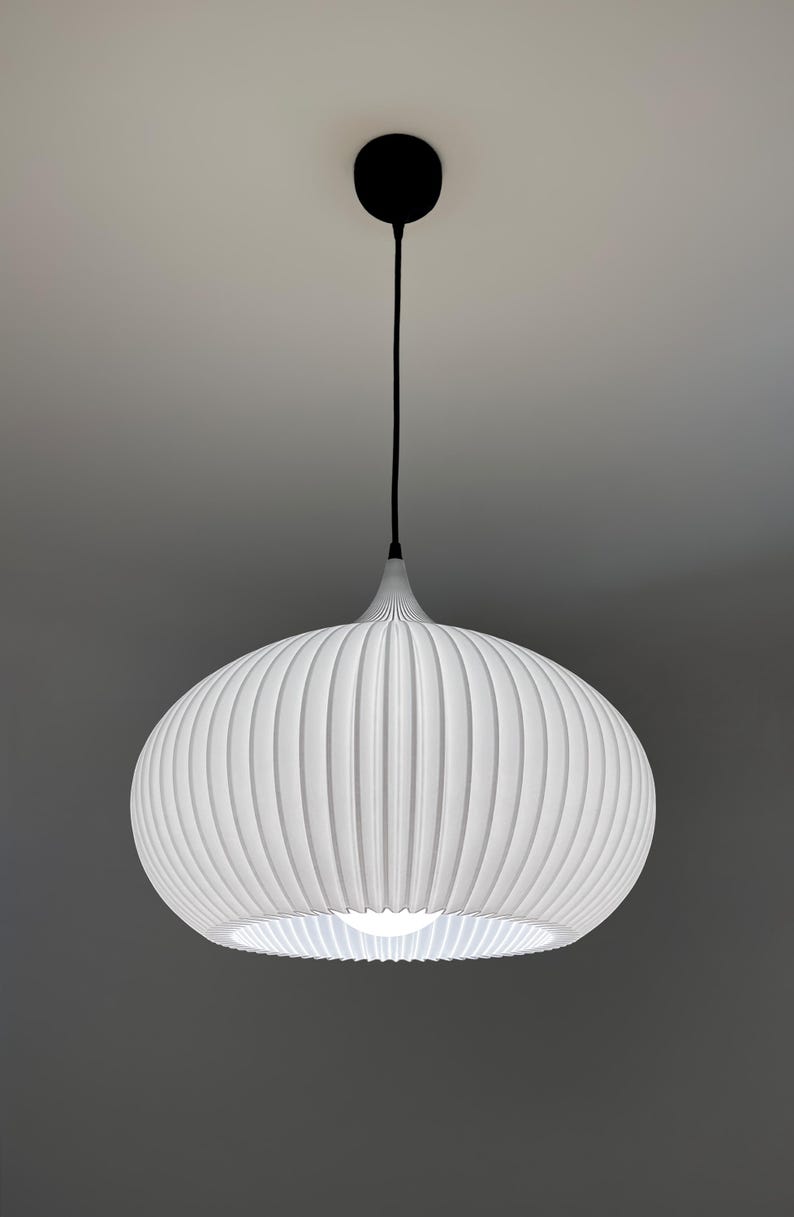A white pendant light fixture with a rounded, ribbed, and pleated shade. The lamp hangs from a black cord attached to a circular black ceiling canopy. A single bright light bulb is visible inside the center of the white shade.