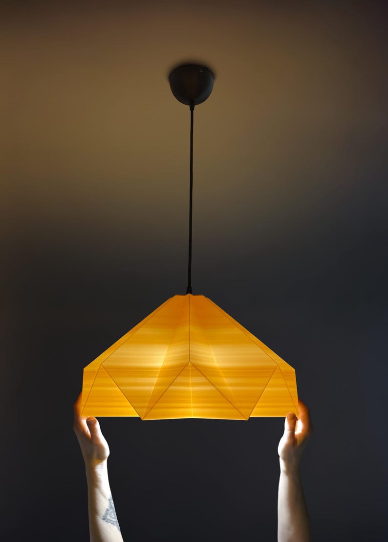 May include: A person holds up a yellow origami-style pendant light with a black cord.