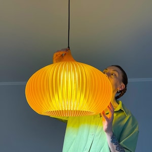 Peut inclure: Une suspension lumineuse jaune vif avec une forme arrondie, nervur&eacute;e et semblable &agrave; une citrouille. La lampe pr&eacute;sente une s&eacute;rie de lattes verticales qui cr&eacute;ent un aspect textur&eacute; et lumineux lorsqu'elle est allum&eacute;e, suspendue par un cordon noir.