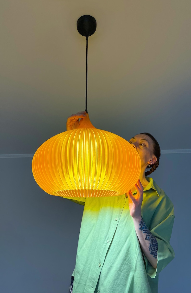 Peut inclure: Une suspension lumineuse jaune vif avec une forme arrondie, nervur&eacute;e et semblable &agrave; une citrouille. La lampe pr&eacute;sente une s&eacute;rie de lattes verticales qui cr&eacute;ent un aspect textur&eacute; et lumineux lorsqu'elle est allum&eacute;e, suspendue par un cordon noir.