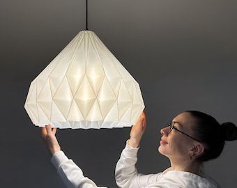 May include: A white geometric pendant lamp with a faceted, diamond-like structure. The shade is made of a translucent material that glows when illuminated, suspended from the ceiling by a thin black cord and a matching black canopy.