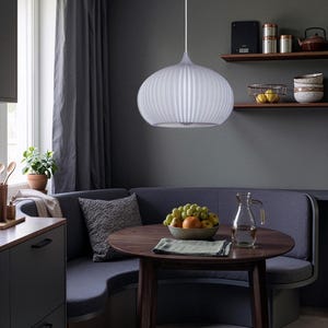 A white, ribbed, spherical pendant light fixture hangs above a round, dark wood dining table. The table holds a bowl of mixed fruit and a glass pitcher. A curved, dark gray upholstered bench seat is positioned behind the table.