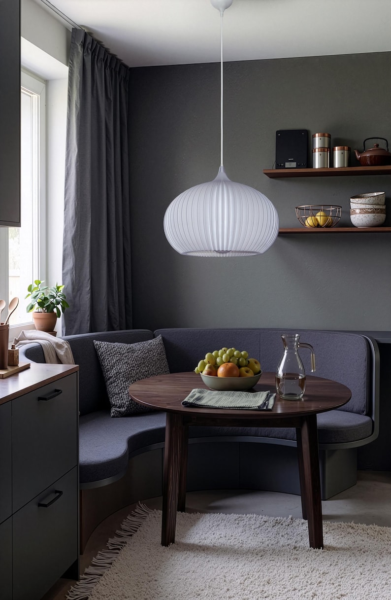 A white, ribbed, spherical pendant light fixture hangs above a round, dark wood dining table. The table holds a bowl of mixed fruit and a glass pitcher. A curved, dark gray upholstered bench seat is positioned behind the table.