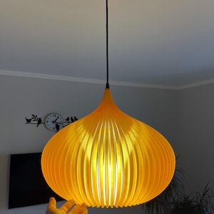 Peut inclure: Une suspension lumineuse jaune vif avec une silhouette arrondie en forme d'oignon et des d&eacute;tails nervur&eacute;s verticaux. La lampe est suspendue au plafond par un fin cordon noir et une fixation circulaire noire assortie au sommet.