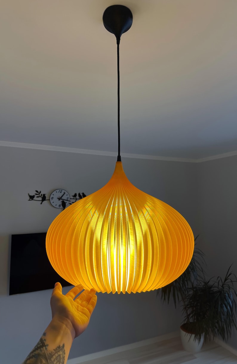 Peut inclure: Une suspension lumineuse jaune vif avec une silhouette arrondie en forme d'oignon et des d&eacute;tails nervur&eacute;s verticaux. La lampe est suspendue au plafond par un fin cordon noir et une fixation circulaire noire assortie au sommet.
