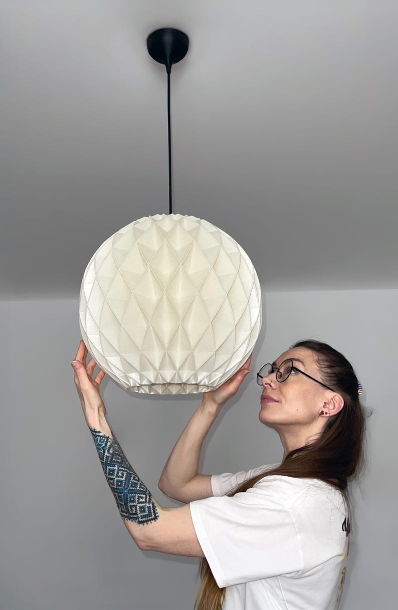 Może przedstawiać: Sferyczna lampa wisząca z białym, składanym, geometrycznym papierowym kloszem o fakturze w romby. Klosz jest zawieszony na suficie za pomocą cienkiego czarnego przewodu przymocowanego do pasującej czarnej okrągłej podsufitki.