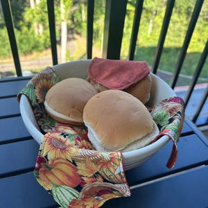 May include: A ceramic bowl filled with hamburger buns, lined with a colorful fabric featuring sunflowers, pumpkins, and autumn leaves. A folded red cloth sits atop the buns. The scene is set on a dark blue table.