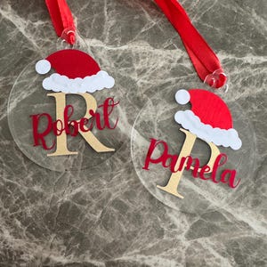 Puede incluir: Dos adornos navideños redondos y transparentes con cintas rojas. Cada adorno presenta un diseño de gorro de Papá Noel, una letra dorada y un nombre en escritura roja. Uno dice "Robert" y el otro "Pamela".
