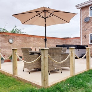 May include: A patio with a wooden deck and a rope railing. There is a table and chairs with a beige umbrella. The patio is surrounded by green grass.