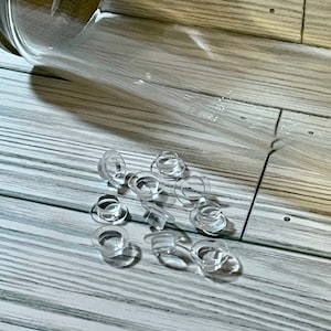 May include: A group of clear plastic eyelets scattered on a light wood surface. The eyelets are small and round, with a hole in the center. A clear glass jar is lying on its side in the background.