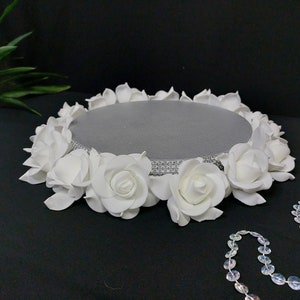 Pode incluir: Um suporte de bolo redondo cinza, decorado com rosas brancas na borda. O suporte tem uma faixa prateada com strass. Uma fileira de contas claras e iridescentes está em primeiro plano. Em uma superfície preta.