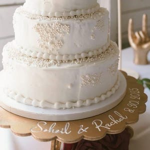 May include: A three-tiered white wedding cake with pearl-like decorations and a gold cake topper that reads "Mr & Mrs Shojai". The cake sits on a gold scalloped cake stand with the names "Scheil & Rachel" and the date "8.30.2025".