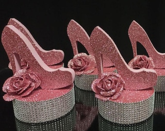May include: Pink glitter high heel shoe decorations with a rose accent. The shoes are set on a silver-colored base. The background is black.