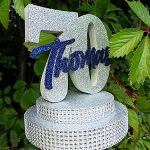 Op de afbeelding: Een zilveren glitter taarttopper in de vorm van het getal 70 met de naam Thomas in donkerblauw glitter-schrift aan de voorzijde. De topper staat op een ronde voet met twee lagen, bedekt met zilveren glitters en rijen strass-steentjes.