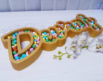 Pode incluir: Decoração de madeira dourada com a palavra «Dream» preenchida com doces coloridos. Orquídeas brancas e vegetação são colocadas perto da decoração sobre uma superfície branca. Os doces são de várias cores.