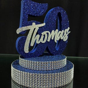 May include: A blue glittery number 50 cake topper featuring the name Thomas in silver script lettering. The base consists of two stacked circular tiers, each wrapped in a band of silver rhinestone mesh, creating a decorative centerpiece.