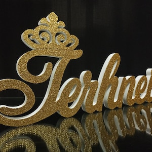 Può includere: Un'opera d'arte con parole ricoperta di glitter dorati che recita "Terhinei" in un carattere corsivo, sormontata da una corona. Le lettere sono bianche con glitter dorati sul davanti. Il pezzo è esposto su una superficie nera riflettente.
