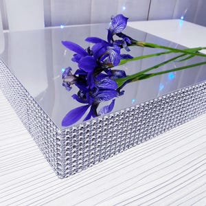 May include: A mirrored, square decorative tray with a sparkling, rhinestone-studded border. The tray reflects a vibrant arrangement of purple iris flowers with green stems. The tray is on a white surface.
