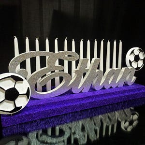 May include: A purple glitter base holds a silver glitter cutout of the name Ethan, flanked by two circular black and white soccer ball icons. A row of thin white candles stands upright behind the name, creating a decorative birthday display.